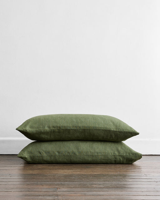 Two Olive Stripe 100% French Flax Linen King Pillowcases by Bed Threads are stacked on a wooden floor against a plain white wall, providing natural, temperature-regulating comfort.
