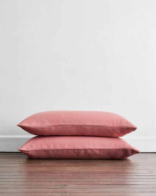 A set of two Pink Clay 100% French Flax Linen King Pillowcases by Bed Threads are stacked on a wooden floor against a plain white wall.