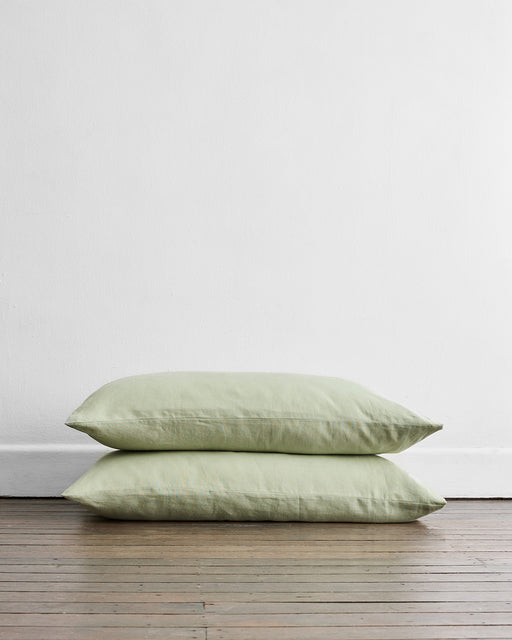 Two Sage 100% French Flax Linen King Pillowcases by Bed Threads are stacked on a wooden floor against a white wall, adding natural elegance and breathability to your space.