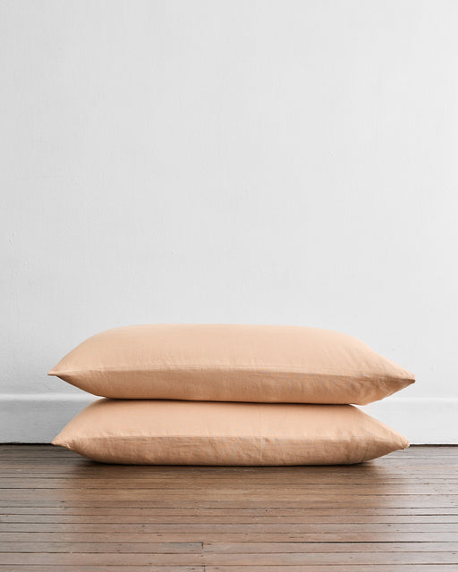 The Bed Threads Terracotta 100% French Flax Linen King Pillowcases (Set of Two) are stacked on a wooden floor against a white wall, offering softness and temperature regulation to your bedroom.