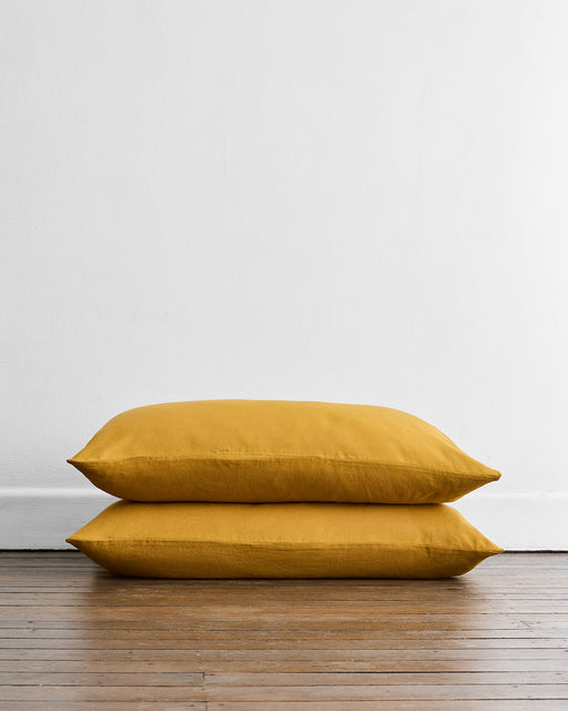 Two Bed Threads Turmeric 100% French Flax Linen King Pillowcases (Set of Two) are stacked on pillows on a wooden floor against a plain white wall.