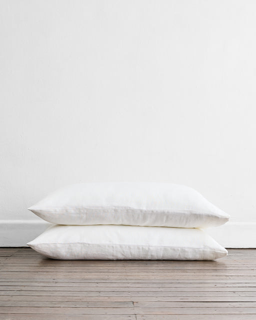 Two Bed Threads White 100% French Flax Linen King Pillowcases (Set of Two) are stacked on a wooden floor against a plain white wall, adding temperature-regulating comfort to your bedding.