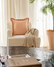 A cream armchair with a Bed Threads Terracotta & Pink Clay French Flax Linen Swirl Cushion Cover and beige throw sits by a large potted plant near sheer curtains. A black table with a book is in front, bathed in soft, natural light. - view