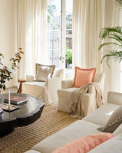 Bright, airy living room with cream armchairs and sofa, pastel throw pillows, Bed Threads’ Oatmeal 100% French Flax Linen Muslin Throw, large windows with sheer curtains, black round coffee table, potted plant, and woven rug for a cozy atmosphere. - view