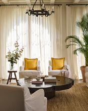 A cozy living room with two white armchairs, each featuring a yellow pillow and a Bed Threads Turmeric & Crème 100% French Flax Linen Swirl Cushion Cover, rustic round wooden coffee table, potted plants, and sheer white curtains with natural light. - view
