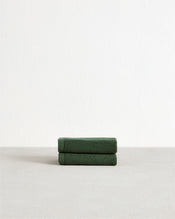 The Bed Threads Moss 100% Cotton Terry Face Washers (Set of Two) are soft, absorbent dark green towels, neatly folded on a light grey surface against a white wall—ideal for daily use. - view