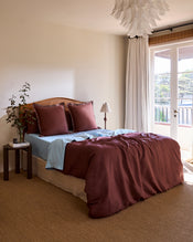 A cozy bedroom with a neatly made bed featuring Bed Threads' Cacao & Coast 100% French Flax Linen Swirl European Pillowcases, a blue sheet, a bedside table with a plant and books, a floor lamp, and large windows adorned with white curtains. - view