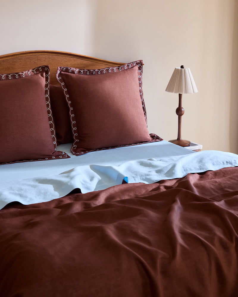 A neatly made bed with Bed Threads Cacao & Coast 100% French Flax Linen Swirl European Pillowcases, light blue French flax linen sheets (170GSM), a brown bedspread, and a small bedside table holding a brown lamp and book.
