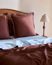 A neatly made bed features Bed Threads’ Cacao & Coast 100% French Flax Linen Swirl European Pillowcases (Set of Two) with decorative edging, a light blue linen sheet, a brown blanket, and a small lamp on the wooden nightstand. - view