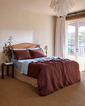 A cozy bedroom features a wooden headboard, blue sheets, maroon bedding, and two Bed Threads Cacao & Coast 100% French Flax Linen Swirl Pillowcases. A bedside table with a plant and lamp is by a large window with white curtains overlooking the balcony. Carpeted floor. - view