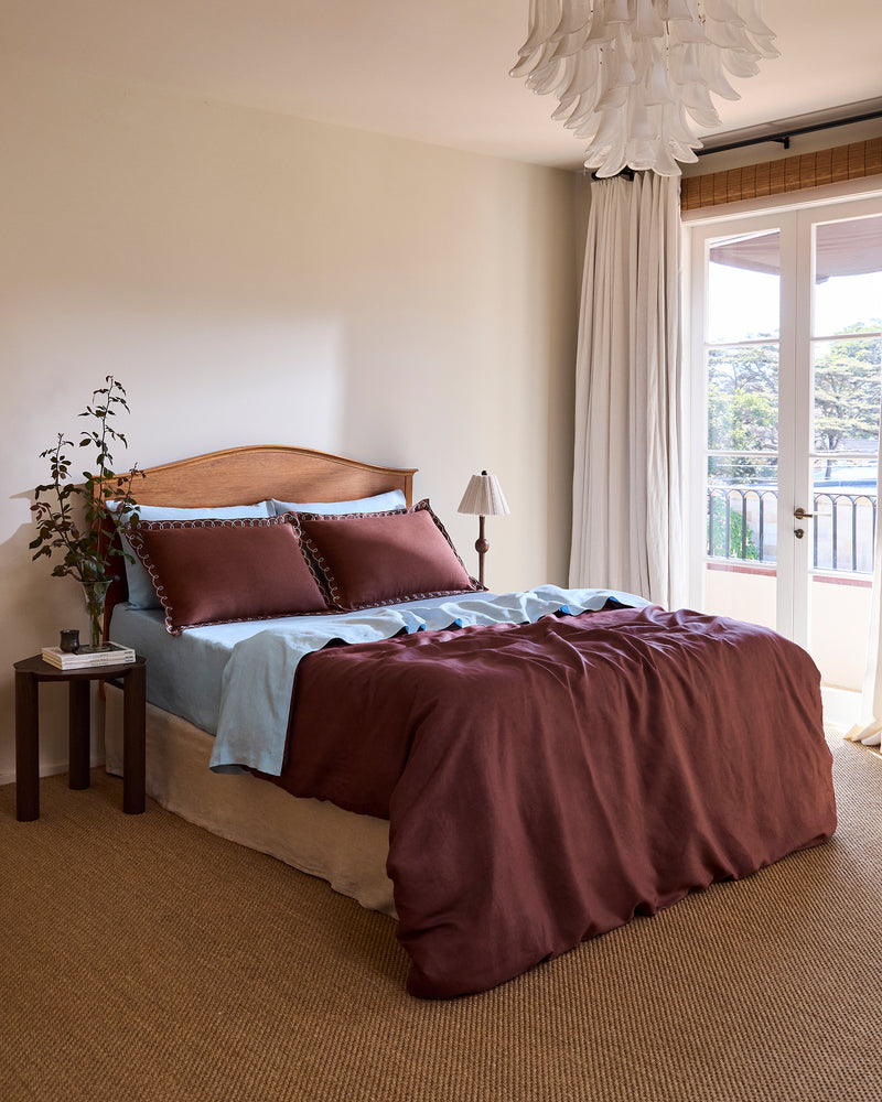 A cozy bedroom features a wooden headboard, blue sheets, maroon bedding, and two Bed Threads Cacao & Coast 100% French Flax Linen Swirl Pillowcases. A bedside table with a plant and lamp is by a large window with white curtains overlooking the balcony. Carpeted floor.