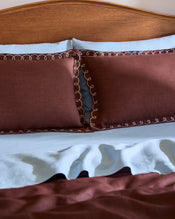 A neatly made bed with light blue sheets and Bed Threads’ Cacao & Coast 100% French Flax Linen Swirl Pillowcases, featuring brown embroidery and white loops, sits against a wooden headboard. The linen bedspread has a brown section at the bottom. - view