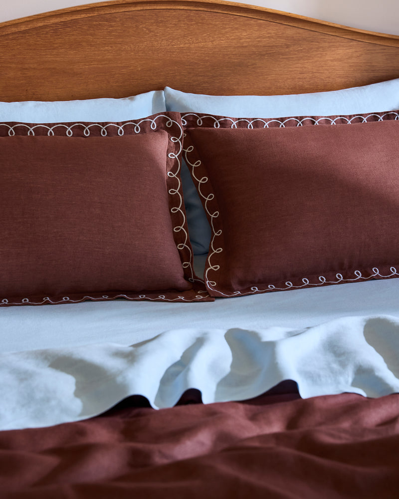 A neatly made bed with light blue sheets and Bed Threads’ Cacao & Coast 100% French Flax Linen Swirl Pillowcases, featuring brown embroidery and white loops, sits against a wooden headboard. The linen bedspread has a brown section at the bottom.