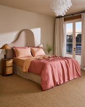 A cozy bedroom with a large bed dressed in Terracotta & Pink Clay 100% French Flax Linen Swirl Pillowcases by Bed Threads, a unique curved headboard, neutral walls, tall curtained windows overlooking trees, and a wicker nightstand with lamp. - view