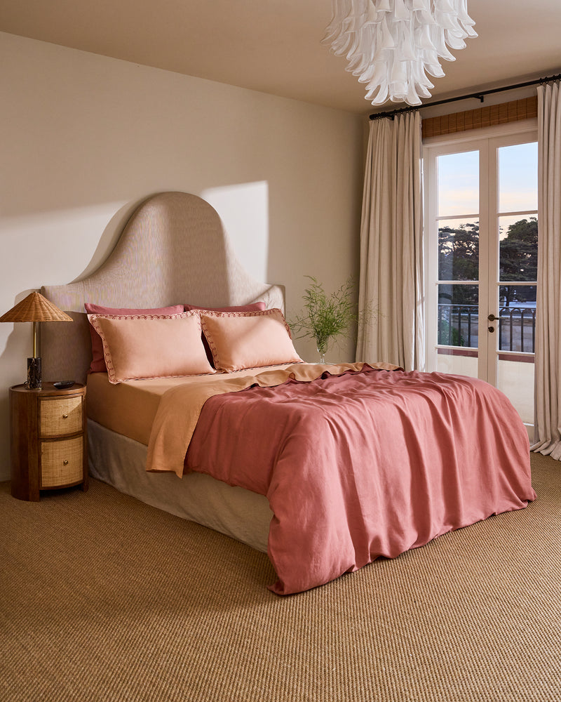 A cozy bedroom with a large bed dressed in Terracotta & Pink Clay 100% French Flax Linen Swirl Pillowcases by Bed Threads, a unique curved headboard, neutral walls, tall curtained windows overlooking trees, and a wicker nightstand with lamp.