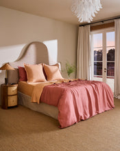 A cozy bedroom features a uniquely shaped upholstered headboard, Bed Threads’ Terracotta & Pink Clay 100% French Flax Linen Swirl European Pillowcases, a wooden nightstand with a lamp, sheer curtains, a glass door, and a modern chandelier. - view