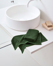 A modern white bathroom sink features a folded dark green towel and Bed Threads Moss 100% Cotton Terry Face Washers (Set of Two) on the counter, with a beige sponge in a wooden dish creating an inviting daily care space. - view