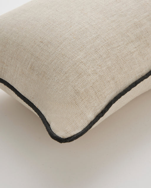 A close-up of the Bed Threads Oatmeal & Charcoal 100% French Flax Linen Piped Lumbar Cushion Cover, featuring black piping along the edge, displayed on a white surface.
