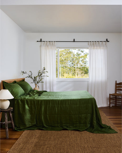 A neatly made bed features the Bed Threads Olive Quilt and matching pillows, all reversible, beside a wooden headboard. A beige vase with leafy branches on a side table near a white curtain completes the calm, minimal bedroom scene.
