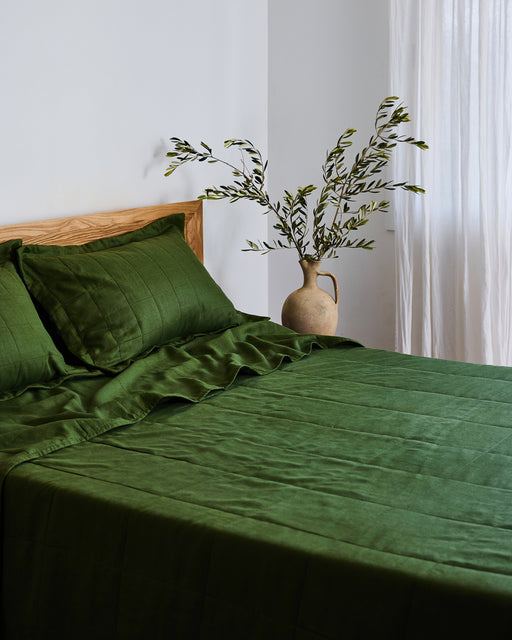 A bed features dark green Olive Quilted Pillowcases (Set of Two) by Bed Threads, paired with a wooden headboard. A beige vase with leafy branches sits on a bedside table beside a sheer white curtain.