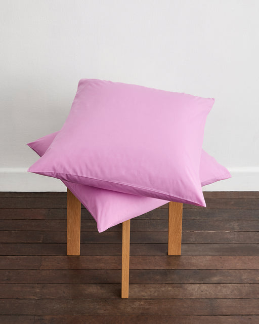 Two light pink Bubblegum 100% Organic Cotton European Pillowcases by Bed Threads are stacked on a small four-legged wooden stool, set on a dark wood floor against a white wall.