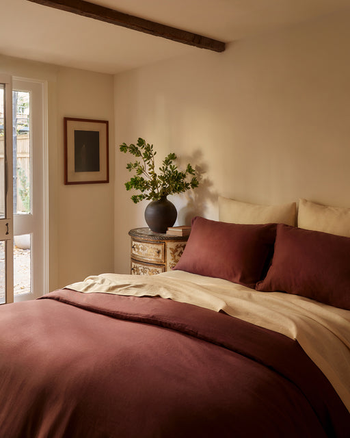 A cozy bedroom features a bed dressed in the Bed Threads Cacao & Crème 100% French Flax Linen Bedding Bundle, a vintage dresser with a dark vase of green branches, framed wall art, and French doors streaming in natural light.