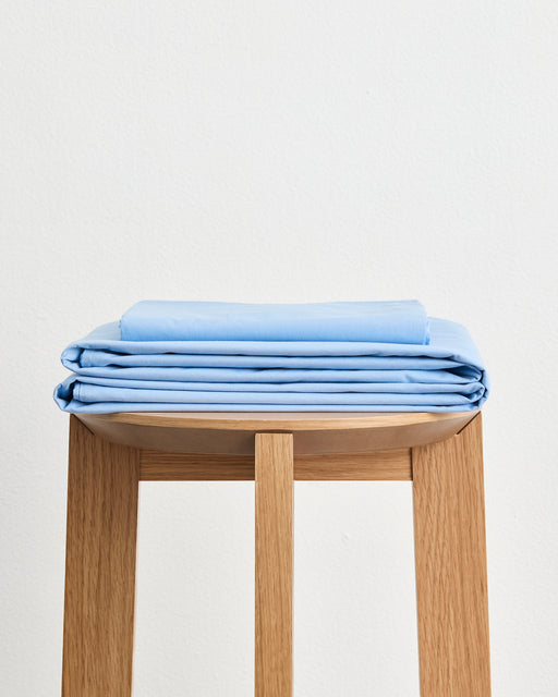 A neatly made bed features the Bed Threads Sky Organic Cotton Flat Sheet, GOTS certified, in light blue with two matching pillows against a white background. Slight wrinkles on the sheet add a relaxed, comfortable look.