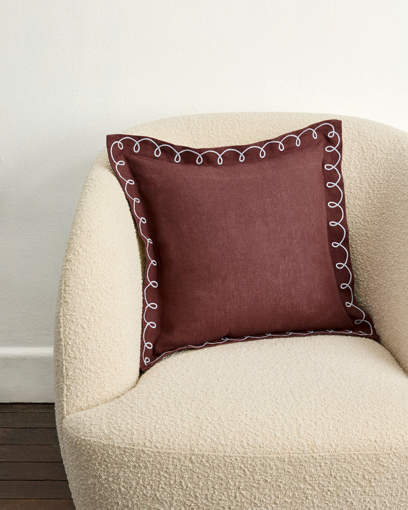 A cream-colored armchair with a square maroon pillow featuring the Bed Threads Cacao & Coast 100% French Flax Linen Swirl Cushion Cover, decorated with white swirl embroidery, sits by a white wall and dark wooden floor.