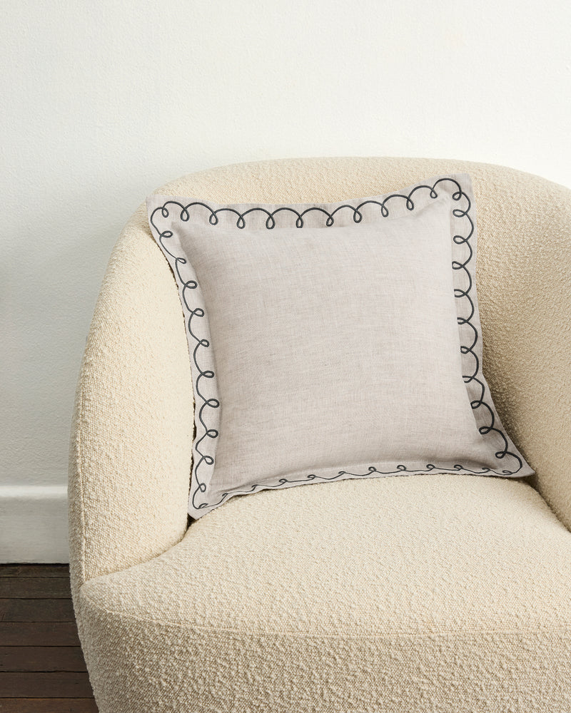 A cream textured armchair with a swirl edge is paired with the Bed Threads Oatmeal & Charcoal 100% French Flax Linen Swirl Cushion Cover, showcasing a black embroidered border, against a white wall and dark wood floor.