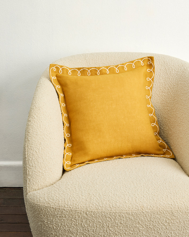 A Turmeric & Crème 100% French Flax Linen Swirl Cushion Cover by Bed Threads sits on a cream-colored textured armchair against a plain white wall.