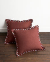 Two square Cacao & Coast 100% French Flax Linen Swirl European Pillowcases by Bed Threads, featuring white decorative edging, rest on a wooden floor against a white wall. - view