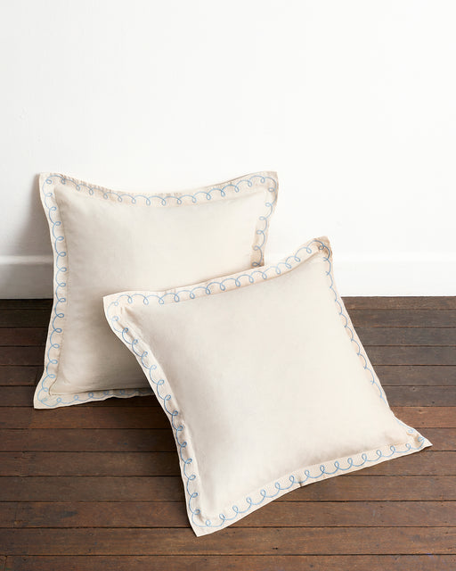 Two Crème & Coast 100% French Flax Linen Swirl European Pillowcases by Bed Threads, set of two, in beige with light blue decorative edge stitching and zip closure, displayed on a wooden floor against a white wall.