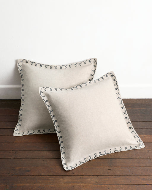 A pair of Bed Threads Oatmeal & Charcoal 100% French Flax Linen Swirl European Pillowcases are styled on a wooden floor against a white wall, highlighting their elegant stitching and classic design.