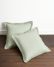 Two Sage & Olive 100% French Flax Linen Swirl European Pillowcases by Bed Threads are arranged on a wooden floor against a white wall—one upright, one flat—showcasing their stylish hue and craftsmanship. - view