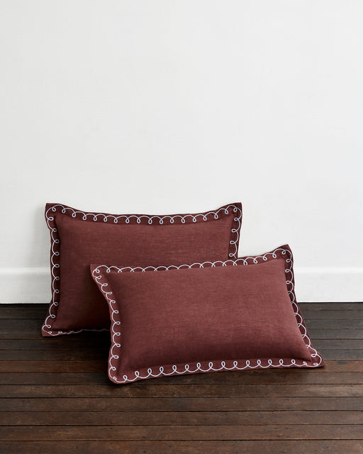 Two Cacao & Coast 100% French Flax Linen Swirl Pillowcases by Bed Threads in maroon, with white decorative stitching, are placed on a dark wooden floor against a plain white wall.