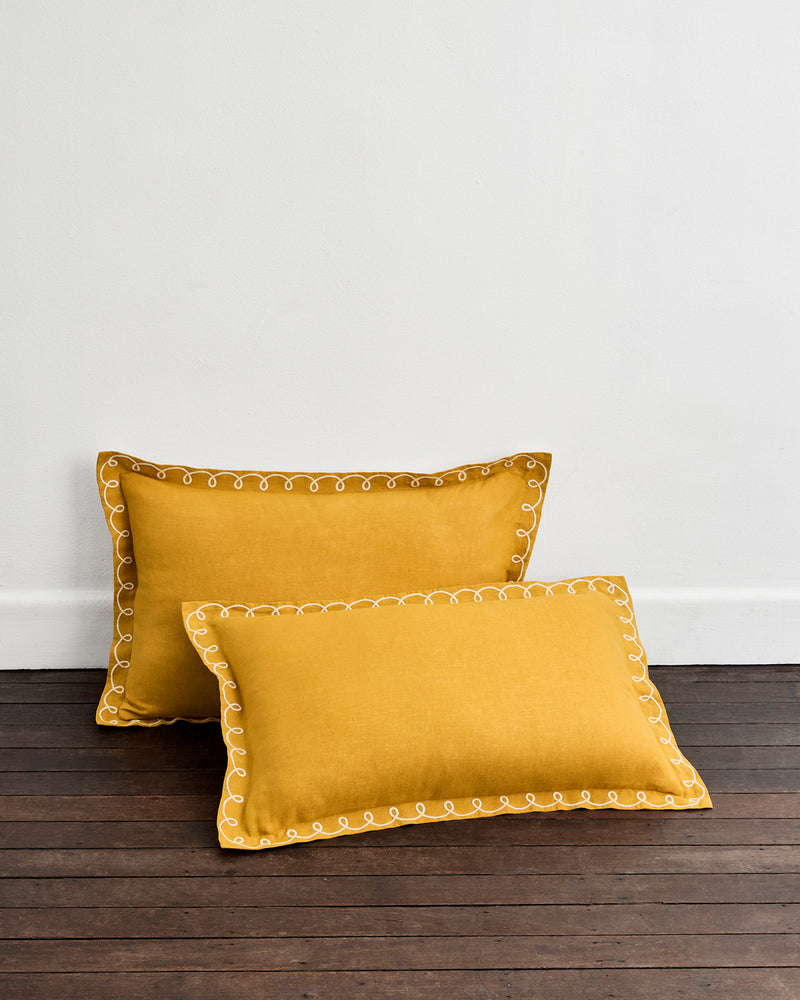 Two Turmeric & Crème 100% French Flax Linen Swirl Pillowcases by Bed Threads are placed on a dark wooden floor, leaning against a white wall.