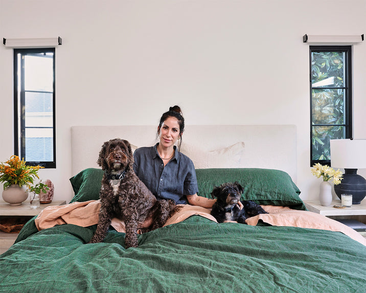 A woman sits on a bed with green covers, smiling, with a large brown dog and a small black dog beside her. There are flowers and decor on nightstands next to windows on each side of the bed.