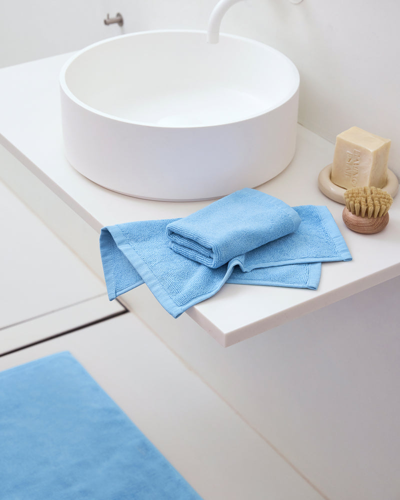 A modern white bathroom sink with a round basin features a bar of soap, scrubbing brush, and neatly folded Bed Threads Sky 100% Cotton Terry Face Washers (Set of Two) for daily care. A matching absorbent terry blue bath mat is on the floor.