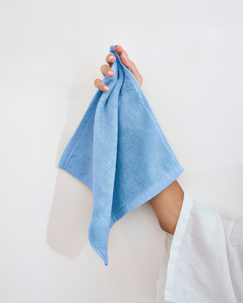 A hand holds a Sky 100% Cotton Terry Face Washer from Bed Threads (Set of Two) against a plain white background. The person, in a white long-sleeve top, shows how these absorbent washers suit daily care routines.