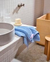 A Bed Threads Sky 100% Cotton Terry Hand Towel, a bar of soap on a dish, and a glass of water sit on a white tiled bathroom sink beside a round basin, with a wooden stool and tub in the background. - view