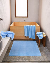A minimalist bathroom with a wooden soaking tub showcases the Sky 100% Cotton Terry Bath Towel Bundle by Bed Threads draped over the tub and stool, a blue bath mat on concrete floors, white tiled walls, and natural light from a small window. - view