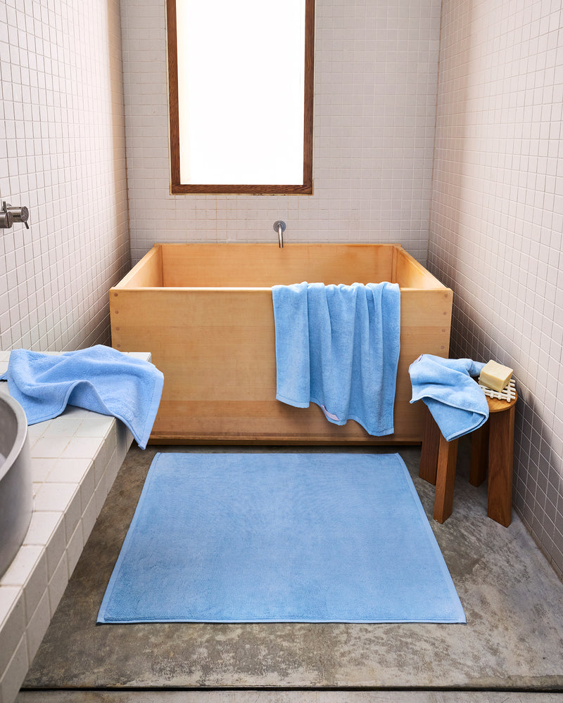 A minimalist bathroom with a wooden soaking tub showcases the Sky 100% Cotton Terry Bath Towel Bundle by Bed Threads draped over the tub and stool, a blue bath mat on concrete floors, white tiled walls, and natural light from a small window.
