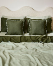 A neatly made bed features a scalloped cream headboard, green French flax linen bedding, and two Bed Threads Olive & Sage 100% French Flax Linen Arch Cushion Covers with white scalloped trim in front of matching shams. - view