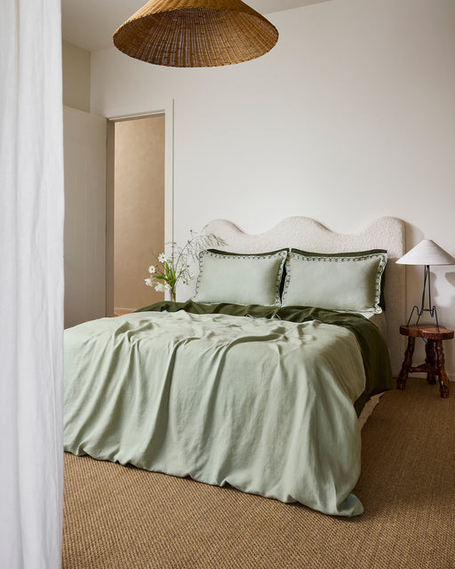 A cozy bedroom features a wavy cream headboard, sage green bedding, and Bed Threads' Sage & Olive 100% French Flax Linen Swirl Pillowcases. Two pillows, a wooden side table with white lamp, woven pendant, natural fiber carpet, and flowers complete the look.