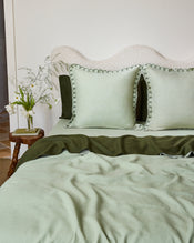 A neatly made bed features Bed Threads’ Sage & Olive 100% French Flax Linen Swirl European Pillowcases (Set of Two), paired with light green bedding, a textured off-white headboard, and a wooden side table holding a glass vase with white wildflowers. - view