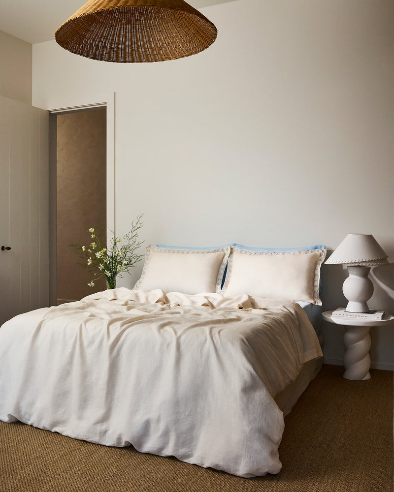 A cozy bedroom with a neatly made bed featuring Bed Threads' Crème & Coast 100% French Flax Linen Swirl Pillowcases (Set of Two), a small white side table with a decorative lamp, a flower vase, and a woven pendant light overhead.