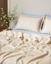 A neatly made bed features Bed Threads' Crème & Coast 100% French Flax Linen Swirl Pillowcases, cream-colored breathable linen bedding, blue-trimmed decorative pillows, a light blue sheet, and a glass vase of white flowers on the bedside table. - view