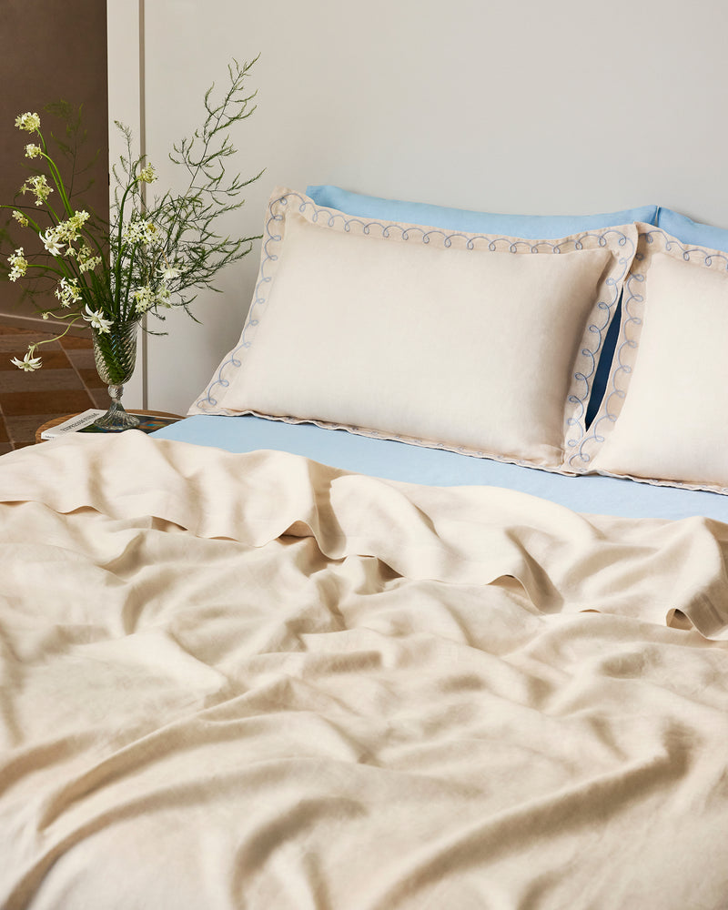 A neatly made bed features Bed Threads' Crème & Coast 100% French Flax Linen Swirl Pillowcases, cream-colored breathable linen bedding, blue-trimmed decorative pillows, a light blue sheet, and a glass vase of white flowers on the bedside table.