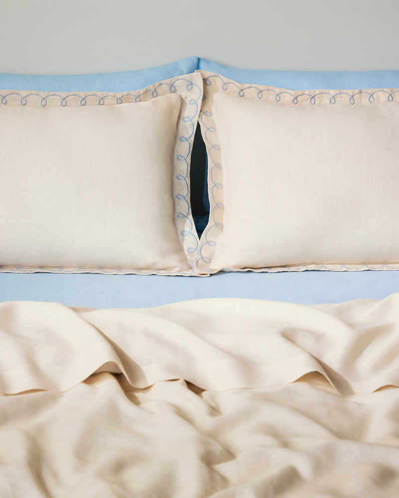 Two Crème & Coast 100% French Flax Linen Swirl Pillowcases by Bed Threads, in cream with blue embroidered edges, rest against a light blue headboard and bedding. The soft, breathable linen appears slightly wrinkled for a relaxed look.