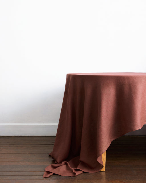 A wooden table draped with the Bed Threads Cacao 100% French Flax Linen Tablecloth sits on a dark wood floor against a plain white wall.